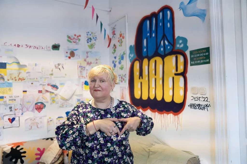 elderly woman holds hands to heart with "No War" spraypainted behind her